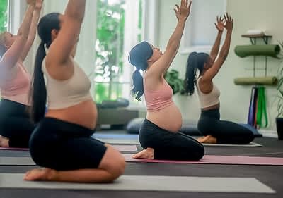 Drei schwangere Frauen sitzen auf Yogamatten in einem hellen Raum und strecken ihre Arme nach oben, während sie Yoga machen, im Hintergrund sind Fenster mit Blick auf grüne Bäume und an der Wand hängen zusammengerollte Yogamatten sowie bunte Yogagurte