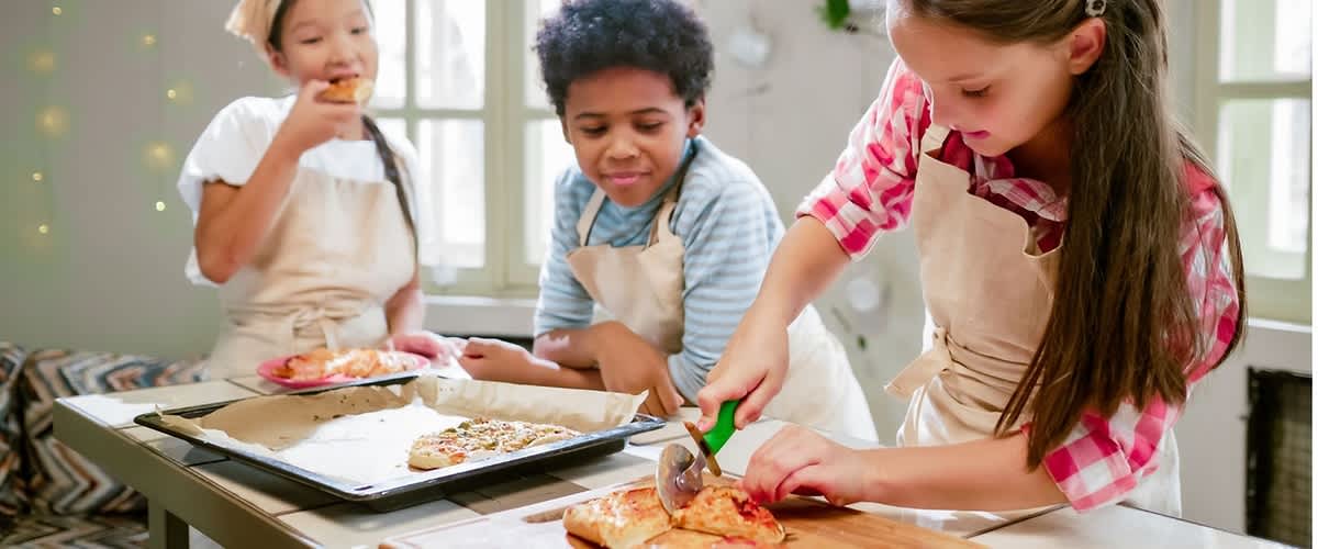Drei Kinder in Schürzen stehen an einem Küchentisch; das Mädchen rechts schneidet mit einem Pizzaroller ein Stück Pizza auf einem Holzbrett, ein Junge in der Mitte und ein weiteres Kind links beobachten und halten jeweils Tellergarnitur, im Hintergrund Fenster mit hellem Tageslicht