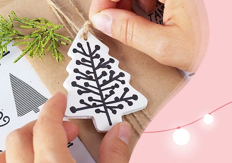 Zwei Hände halten einen weißen Anhänger in Form eines Tannenbaums mit schwarzem Zweigmuster über einem braunen Geschenkpapier mit grünen Tannenzweigen