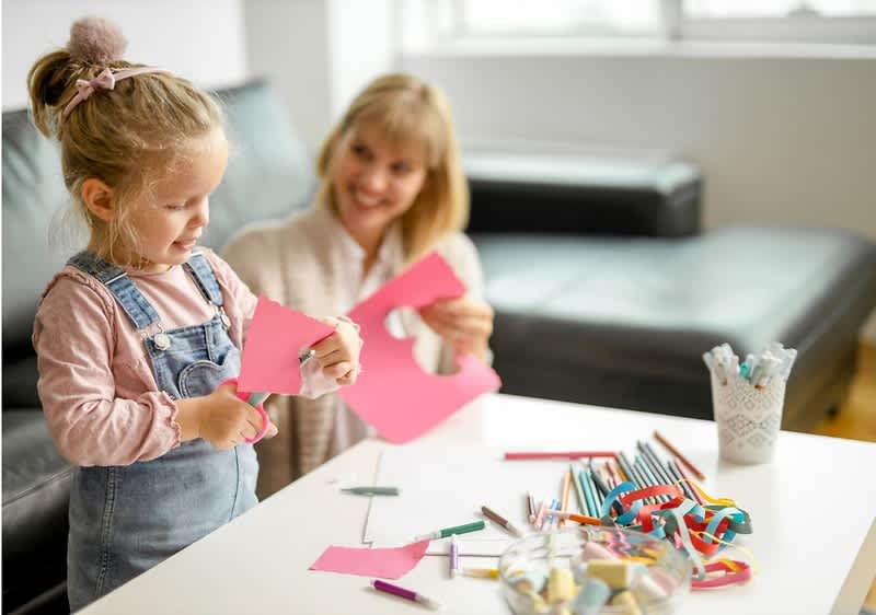 Ein Kind schneidet pinkes Papier mit einer Schere, eine erwachsene Person hält weiteres zugeschnittenes pinkes Papier; auf dem Tisch liegen Buntstifte, Stiftebecher, Papierstreifen und Bastelmaterialien