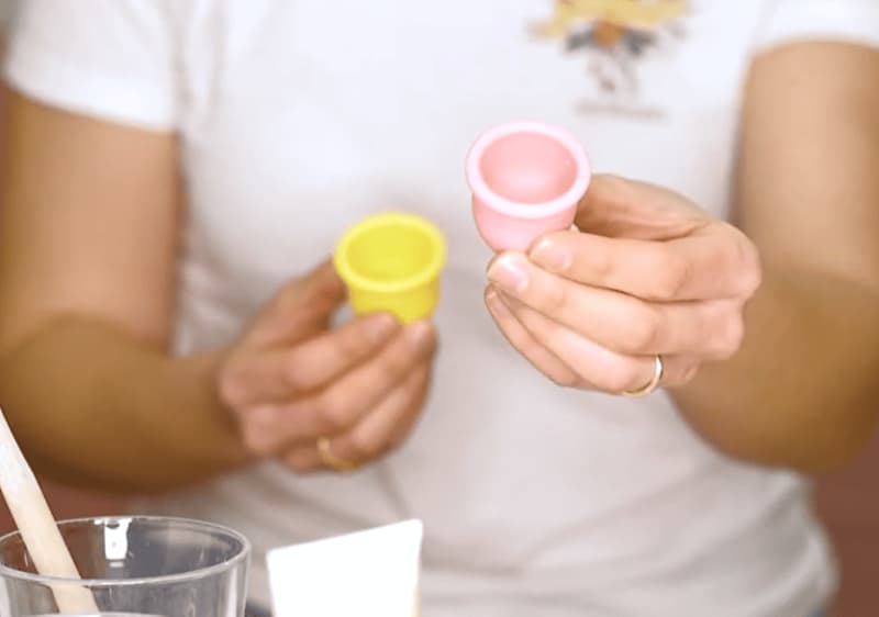 YouTube Video von einhorn zum einsetzen von Menstruationstassen: Person von der Brust abwärts hält in beiden Händen zwei Menstruationstassen, links eine gelbe, rechts eine rosafarbene, im Vordergrund ein Glas mit Holzstäbchen und eine Tubenöffnung sichtbar