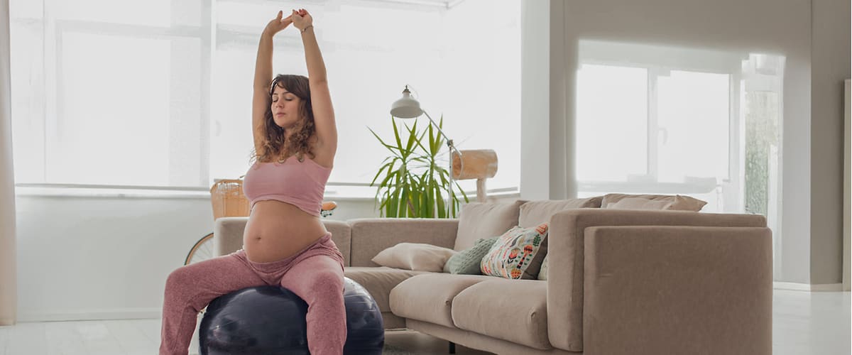 Eine schwangere Frau in rosa Sportkleidung sitzt auf einem Gymnastikball und streckt ihre Arme nach oben, im Hintergrund ein beiges Sofa und eine grüne Pflanze