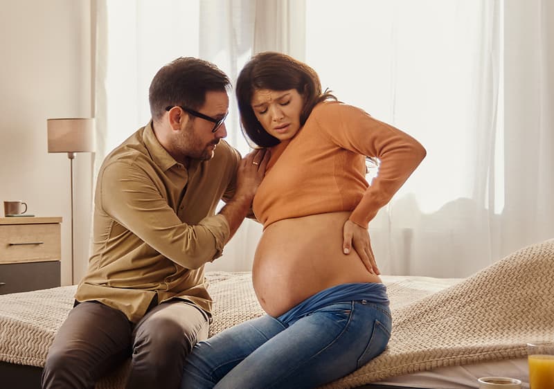 Ein Mann sitzt auf einem Bett und legt seine Hand liebevoll auf den Bauch einer schwangeren Frau, die in einem orangefarbenen Pullover und blauen Jeans gekleidet ist, während sie ihre Hand auf ihren Rücken stützt