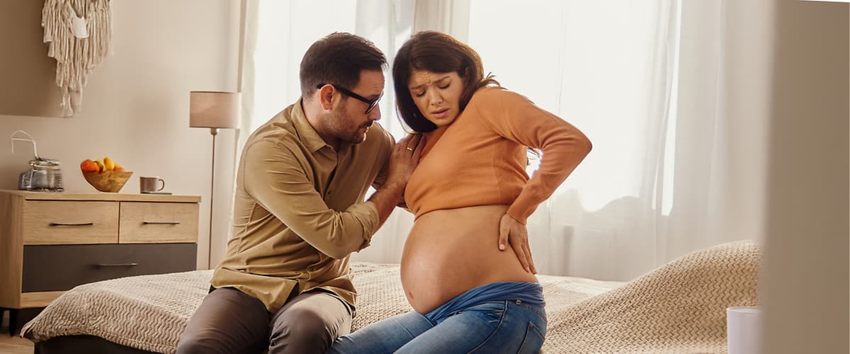 Ein Mann sitzt auf einem Bett und legt seine Hand liebevoll auf den Bauch einer schwangeren Frau, die in einem orangefarbenen Pullover und blauen Jeans gekleidet ist, während sie ihre Hand auf ihren Rücken stützt