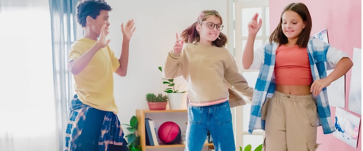 Drei Kinder springen fröhlich auf einem Bett in einem hellen Schlafzimmer, links ein Kind mit gelbem T‑Shirt und dunkler Hose, in der Mitte ein Kind mit beigem Pullover und blauer Jeans, rechts ein Kind mit orangenem Top und karierter Hemdjacke über beigen Cargo‑Hosen, im Hintergrund Pflanzen, ein Regal mit Spielzeug und rosa Wand