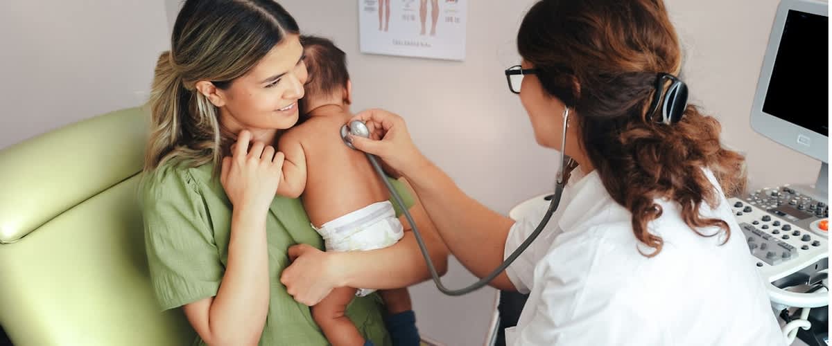 Mutter hält Baby mit Windel auf dem Arm, Ärztin hört mit Stethoskop den Rücken des Babys ab