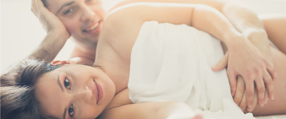 Mann liegt hinter schwangeren Frau, beide haben die Hand auf dem Babybauch