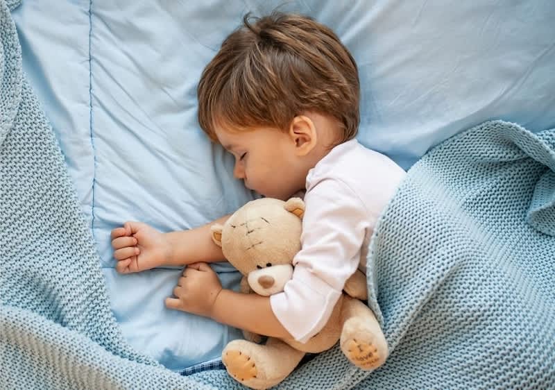 Schlafendes Kleinkind auf der Seite in blauem Bett, umarmt ein braunes Stofftier in Bärform, Decke und Bettwäsche hellblau