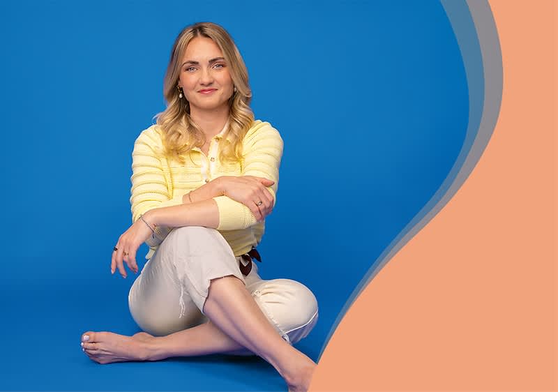 Anne Bäumler sitzt barfuß auf blauem Boden vor blauem Hintergrund, trägt einen gelben Pullover und eine helle Hose, Arme vor dem Oberkörper verschränkt.