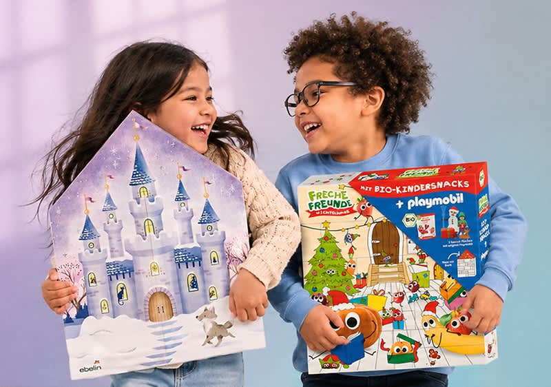 Zwei Kinder stehen nebeneinander, lachen sich an und halten jeweils ein Adventskalender-Türchenbild: links ein großes Haus-förmiges Motiv mit lila Märchenschloss, Schneelandschaft und kleinem Hund, rechts ein bunter rechteckiger Adventskalender mit Weihnachtsbaum, Playmobil- und Tiermotiven sowie der Aufschrift „Freche Freunde“ bzw. „playmobil“. Bild mithilfe von KI erstellt