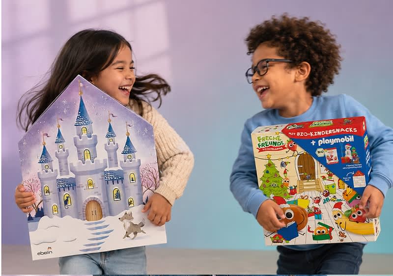 Zwei Kinder stehen nebeneinander und halten jeweils ein Adventskalender-Türchenbild: links ein großes Haus-förmiges Motiv mit lila Märchenschloss, Schneelandschaft und kleinem Hund, rechts ein bunter rechteckiger Adventskalender mit Weihnachtsbaum, Playmobil- und Tiermotiven sowie der Aufschrift „Freche Freunde“ bzw. „playmobil“