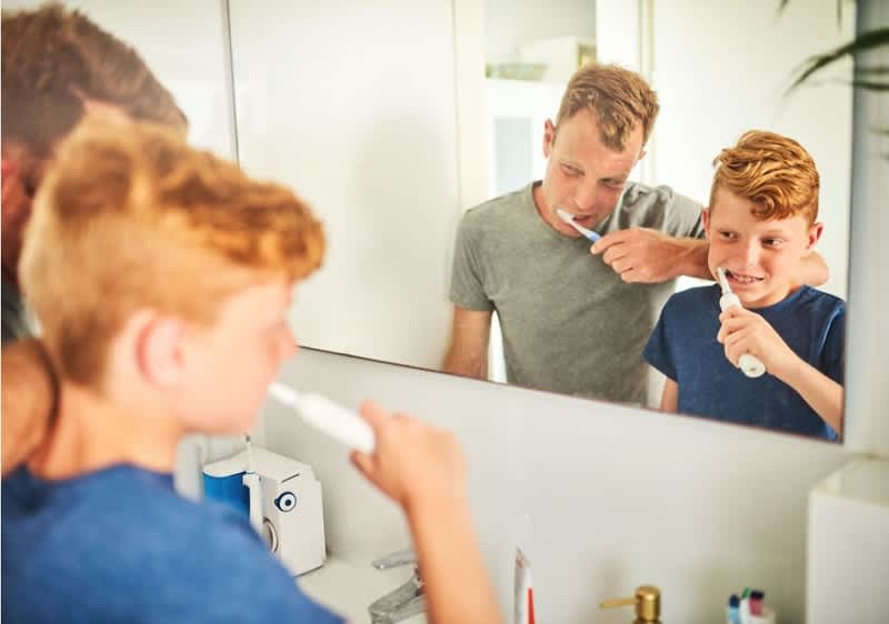 Ein Junge putzt sich die Zähne im Badezimmer, während ein Erwachsener neben ihm steht und in den Spiegel schaut. Im Hintergrund sind ein Zahnputzbecher und ein Wasserhahn zu sehen.