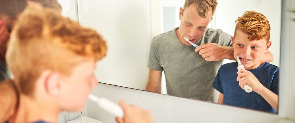 Ein Junge putzt sich die Zähne im Badezimmer, während ein Erwachsener neben ihm steht und in den Spiegel schaut. Im Hintergrund sind ein Zahnputzbecher und ein Wasserhahn zu sehen.