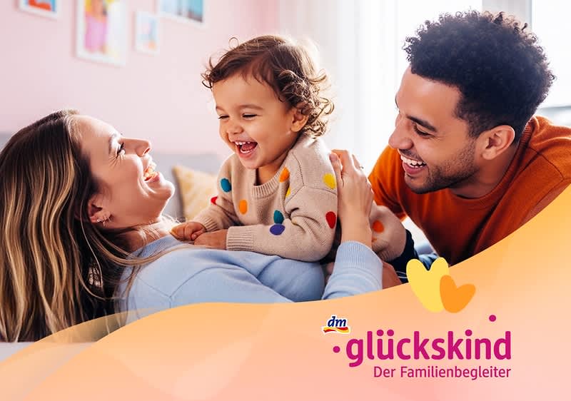 Ein Kleinkind mit einem bunten Pullover sitzt auf dem Bauch einer liegenden Frau, während ein Mann sich zu ihnen beugt. Unten rechts das Logo von dm glückskind, Der Familienbegleiter.. Bild mithilfe von KI erstellt