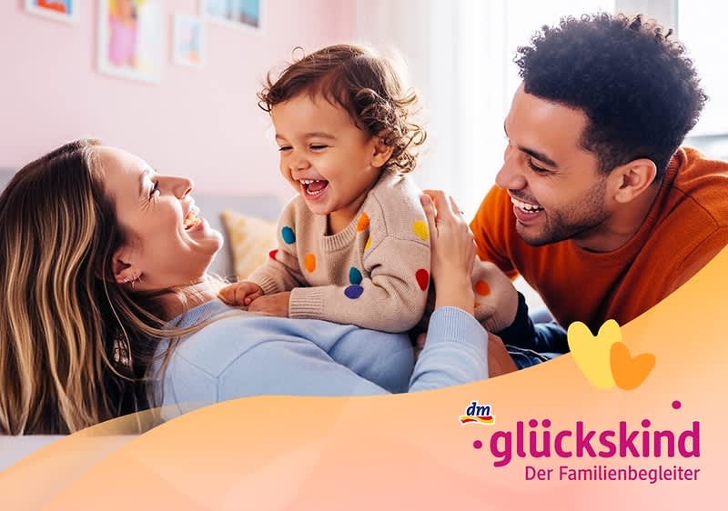 Ein Kleinkind mit einem bunten Pullover sitzt auf dem Bauch einer liegenden Frau, während ein Mann sich zu ihnen beugt. Unten rechts das Logo von dm glückskind, Der Familienbegleiter.