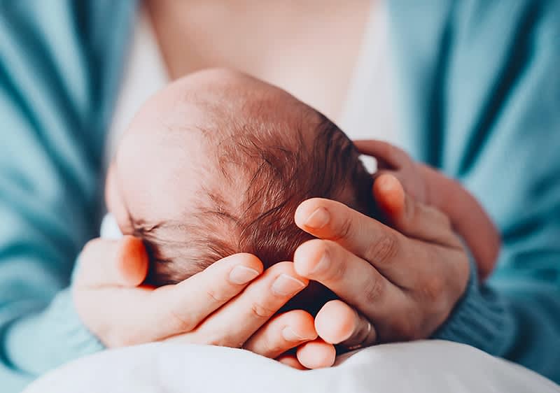 Elternhand hält sanft den Kopf eines neugeborenen Babys, im oberen rechten Bildbereich Logo und Schriftzug dm glückskind