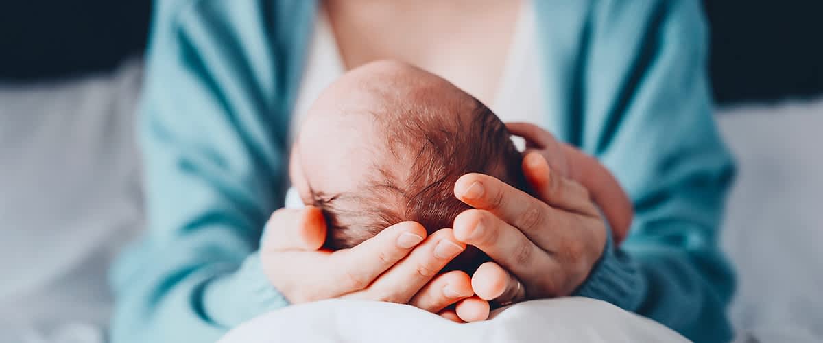 Elternhand hält sanft den Kopf eines neugeborenen Babys, im oberen rechten Bildbereich Logo und Schriftzug dm glückskind