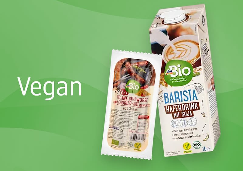 Eine grüne Verpackung von dmBio Barista Haferdrink mit Soja neben einer Packung dmBio Vegane Bratwurst, daneben der weiße Schriftzug „Vegan“ auf grünem Hintergrund
