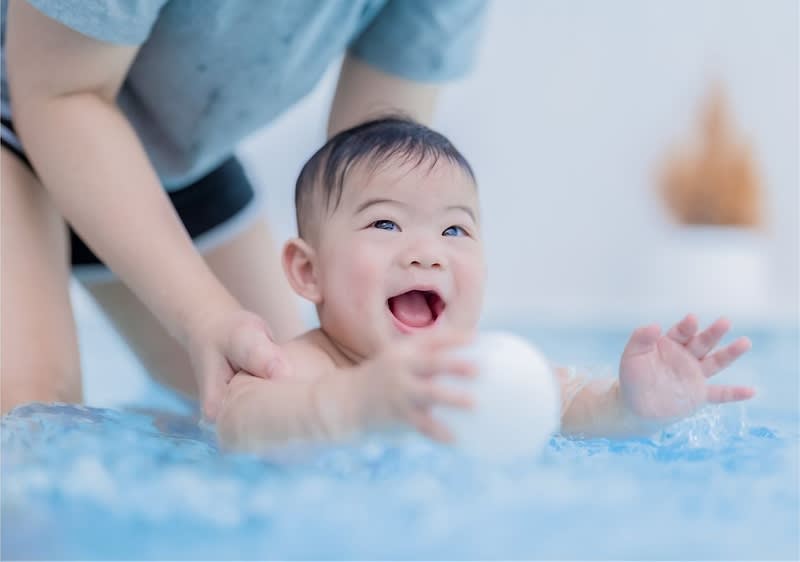 Ein Kleinkind liegt auf dem Bauch auf einer blauen Decke und greift nach einem weißen Spielball, während eine erwachsene Person das Kind sanft an den Seiten stützt, nur Oberkörper der Erwachsenen zu sehen, heller, neutraler Hintergrund