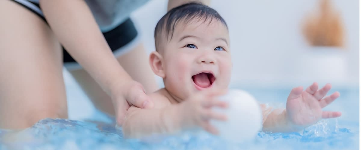 Ein Kleinkind liegt auf dem Bauch auf einer blauen Decke und greift nach einem weißen Spielball, während eine erwachsene Person das Kind sanft an den Seiten stützt, nur Oberkörper der Erwachsenen zu sehen, heller, neutraler Hintergrund