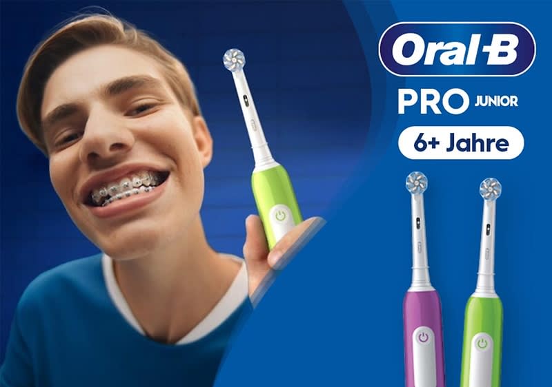 Werbeposter mit dem Logo von Oral‑B oben rechts, Schriftzug PRO JUNIOR und 6+ Jahre, links eine Person mit blauem Oberteil hält eine grün-weiße elektrische Kinderzahnbürste, rechts zwei weitere elektrische Kinderzahnbürsten in Lila-Grün mit runden Bürstenköpfen sichtbar