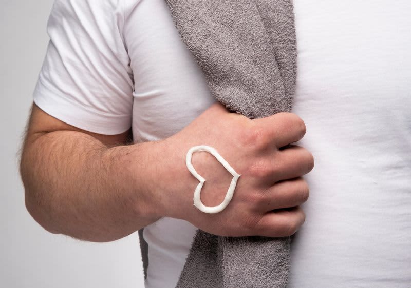 Mann in weißem T-Shirt greift in ein Handtuch, das über seiner Schulter hängt, auf der Hand hat er ein Herz aus Creme.