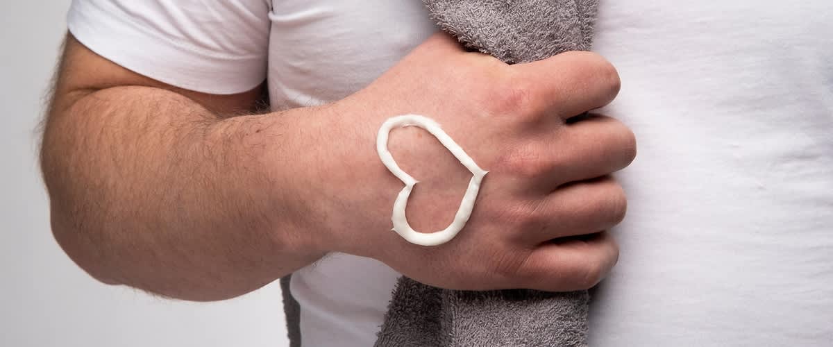 Mann in weißem T-Shirt greift in ein Handtuch, das über seiner Schulter hängt, auf der Hand hat er ein Herz aus Creme.