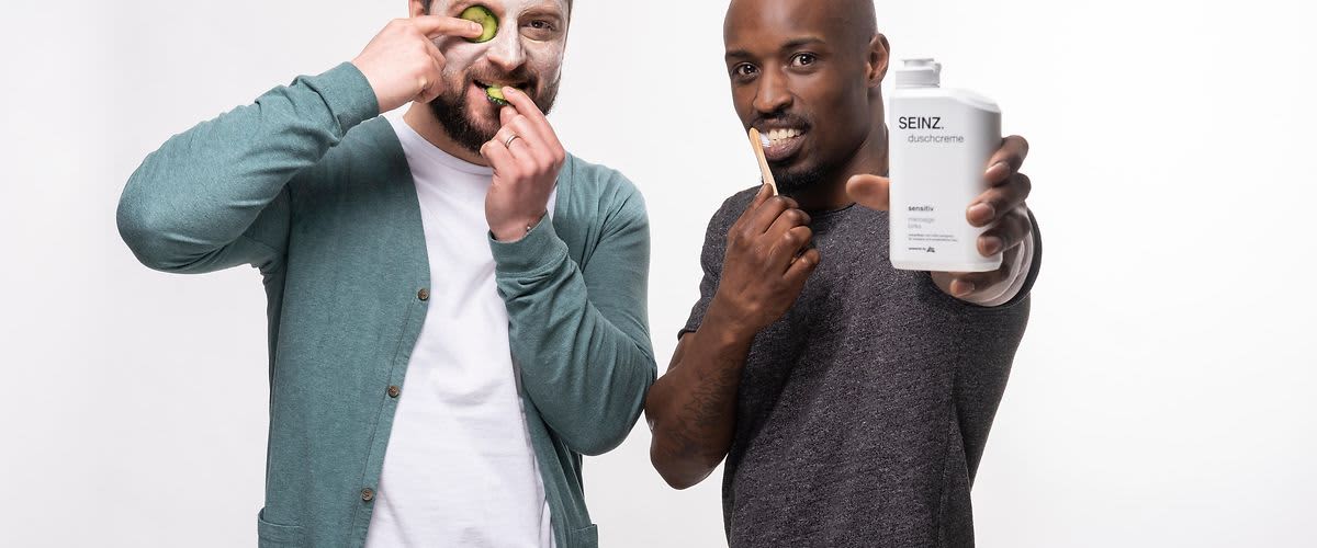 Zwei bärtige Männer, einer mit Gurkenmaske und einer mit Zahnbürste und SEINZ. Duschcreme in der Hand.