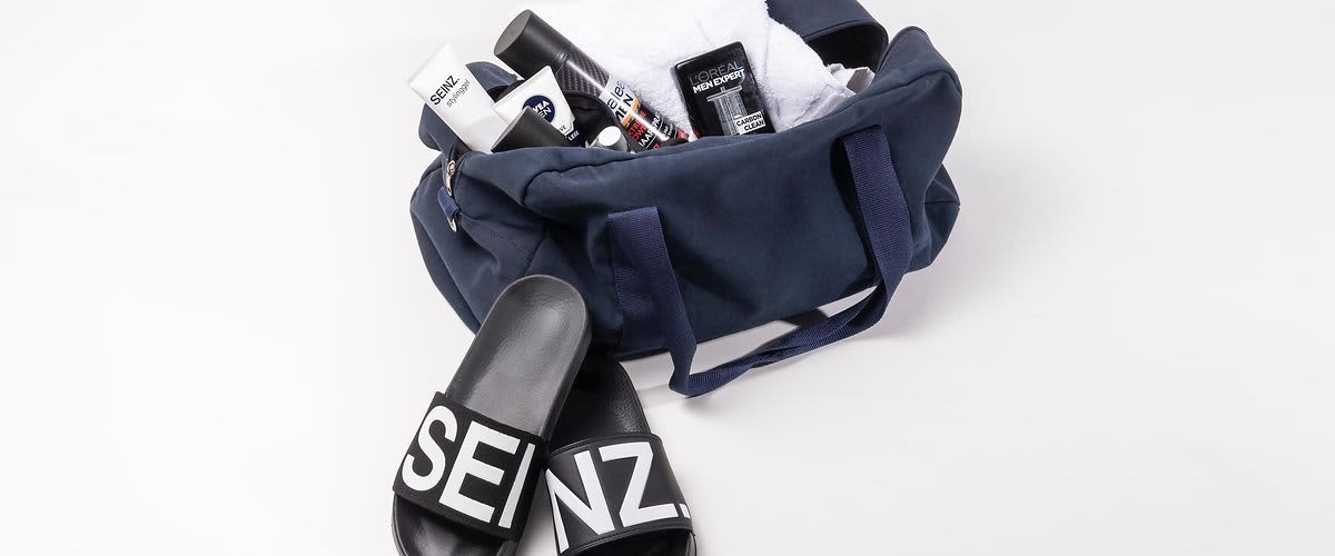 Eine Sporttasche mit Badelatschen von SEINZ. Gefüllt ist die Tasche mit Produkten für die Männerpflege.
