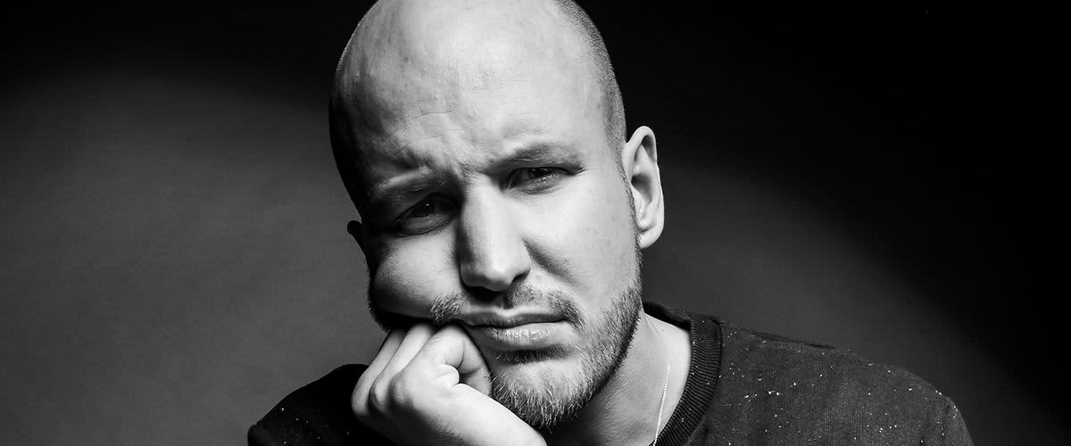 Eine Person in einem dunklen, gepunkteten Oberteil stützt ihren Kopf auf die Hand. Gesicht ist unscharf gemacht. Hintergrund ist dunkel.