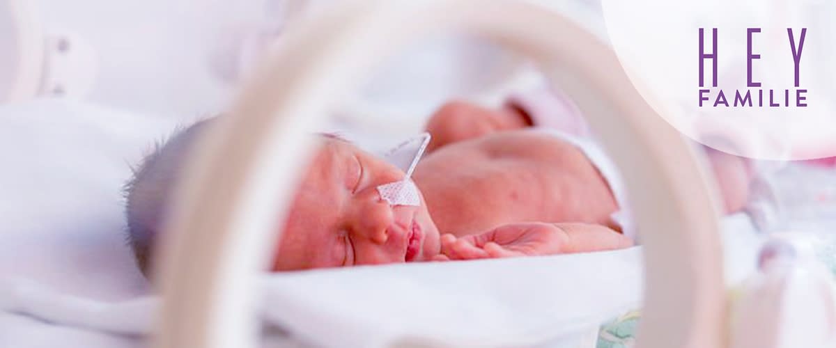 Frühgeborenes Baby mit Nasensonde liegt schlafend in einer Klinik-Inkubator-Umgebung, oben rechts violetter Schriftzug „HEY FAMILIE“