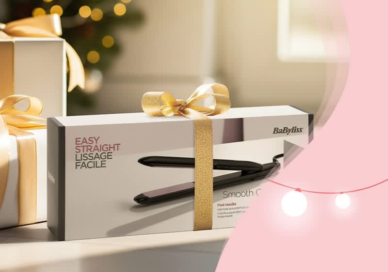 Eine Geschenkbox mit einem Haarglätter von Babyliss, verziert mit einer goldenen Schleife, steht auf einem Tisch. Die Verpackung zeigt den Haarglätter und trägt die Aufschrift „Easy Straight Lissage Facile“