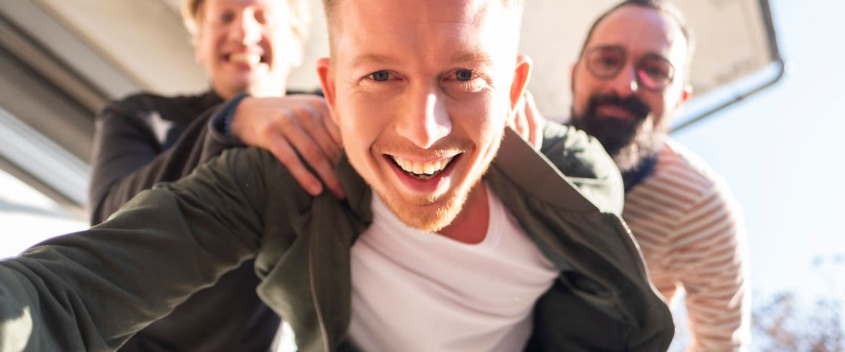 Drei Personen stehen dicht beieinander, eine Person im Vordergrund trägt ein weißes T-Shirt und eine grüne Jacke. Die anderen beiden Personen sind im Hintergrund, eine trägt ein gestreiftes Oberteil