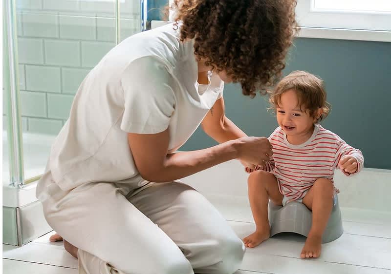 Eine Person mit lockigem Haar und weißem Oberteil kniet auf dem Boden eines Badezimmers und hält die Hände eines Kleinkindes, das auf einem grauen Töpfchen sitzt und einen gestreiften Einteiler trägt, während im Hintergrund eine Duschkabine und ein Fenster zu sehen sind