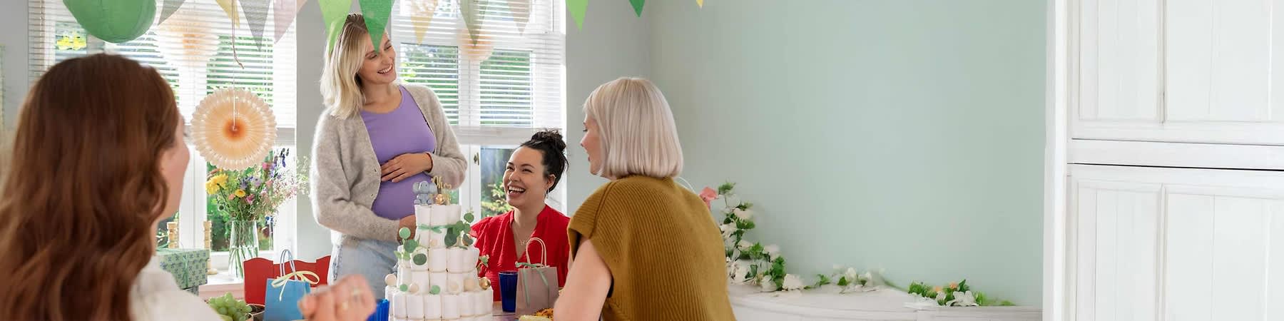 Eine Gruppe von vier Frauen sitzt an einem Tisch in einem hellen Raum, der mit bunten Wimpeln dekoriert ist, auf dem Tisch steht eine Windeltorte mit grünen und weißen Dekorationen, daneben befinden sich Geschenktüten, Blumen und verschiedene Snacks in Schüsseln, während eine Frau in der Mitte steht und ihre Hand auf ihren Bauch legt, die anderen Frauen sitzen um den Tisch und schauen zu ihr hin