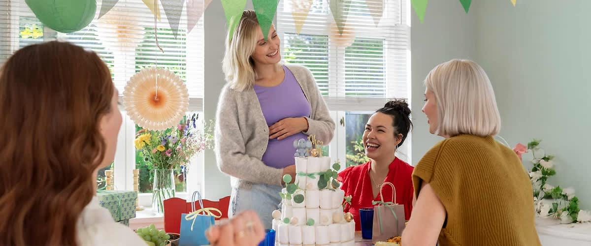 Eine Gruppe von vier Frauen sitzt an einem Tisch in einem hellen Raum, der mit bunten Wimpeln dekoriert ist, auf dem Tisch steht eine Windeltorte mit grünen und weißen Dekorationen, daneben befinden sich Geschenktüten, Blumen und verschiedene Snacks in Schüsseln, während eine Frau in der Mitte steht und ihre Hand auf ihren Bauch legt, die anderen Frauen sitzen um den Tisch und schauen zu ihr hin