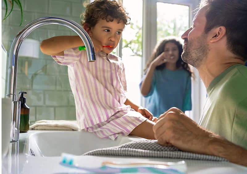 Ein kleines Kind in gestreiftem Schlafanzug sitzt auf einem Waschbecken und putzt sich die Zähne, während ein Mann mit einem Handtuch daneben steht und ihm zusieht, im Hintergrund sind zwei weitere Personen, eine davon ebenfalls mit Zahnbürste, und auf dem Waschbeckenrand liegt eine Zahnbürste neben einer Flüssigseifenspenderflasche.