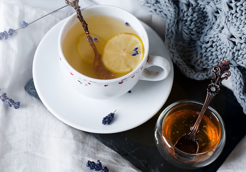 Eine Tasse Tee mit Löffel und Zitronenscheibe. Daneben steht ein Schälchen mit Honig.