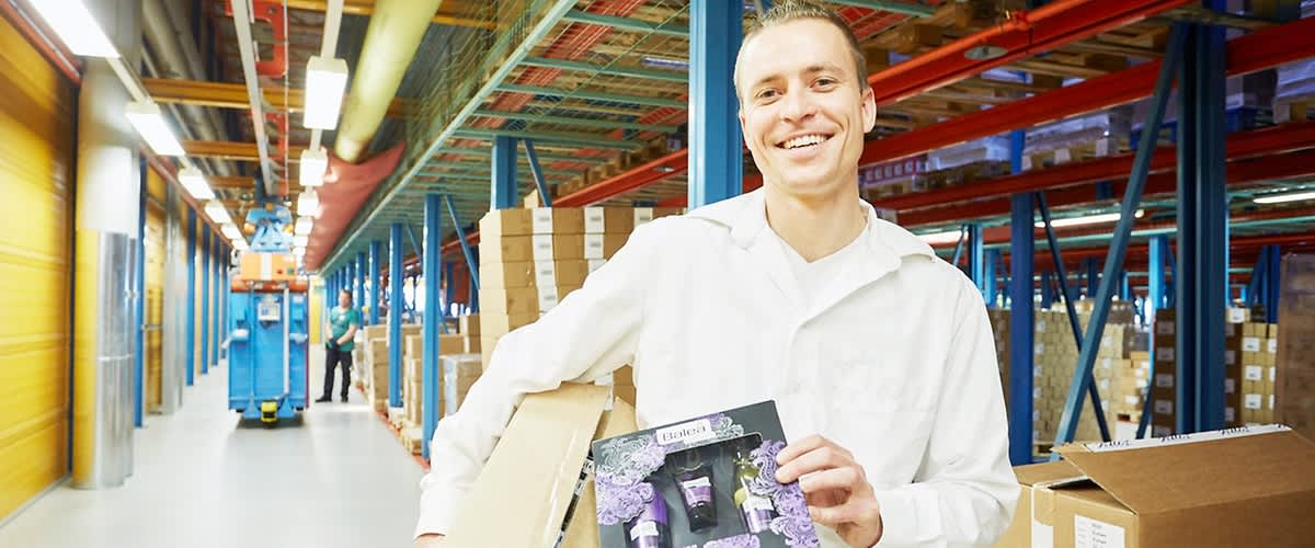 Foto vom dm-Verteilzentrum Weilerswist: Ein Mitarbeiter in einem weißen Kittel steht in einem Lager und hält eine Packung mit lila und schwarzen Grafiken in der Hand, während er gleichzeitig einen großen Karton trägt, im Hintergrund sind Regale mit gestapelten Kartons und eine blaue Hebebühne zu sehen