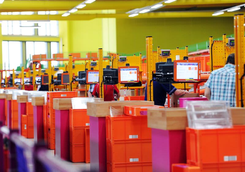 VZ Wustermark: Mehrere Personen arbeiten an orangefarbenen Arbeitsstationen in einem großen, hell beleuchteten Raum mit gelben Wänden und Decken, jede Station ist mit einem Computerbildschirm ausgestattet und die Arbeitsflächen sind mit Holzelementen versehen, im Hintergrund sind große Fenster zu sehen, die zusätzliches Licht in den Raum lassen
