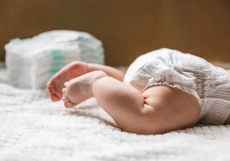 Beinchen und Füße eines liegenden Babys in einer Einwegwindel, das auf einer weißen, strukturierten Decke liegt, im Hintergrund ein Stapel weiterer Windeln ohne erkennbare Markenzeichen