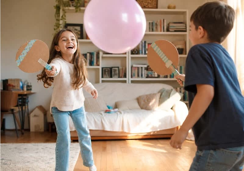Zwei Kinder in einem Wohnzimmer schlagen mit selbstgebastelten Paddeln aus Karton einen großen rosa Ball hin und her