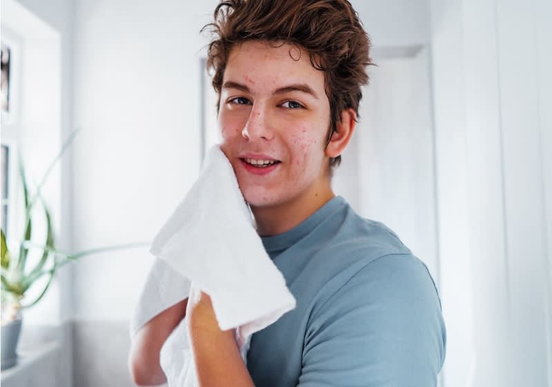 junger Mensch in blauem T‑Shirt trocknet sich mit einem weißen Handtuch im Bad, im Hintergrund Fenster mit Pflanze