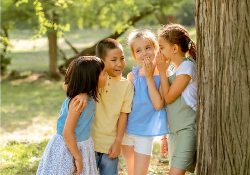 Vier Kinder stehen dicht beieinander an einem Baum im Park; zwei flüstern, zwei hören zu, alle in sommerlicher Freizeitkleidung, helles Sonnenlicht im Hintergrund