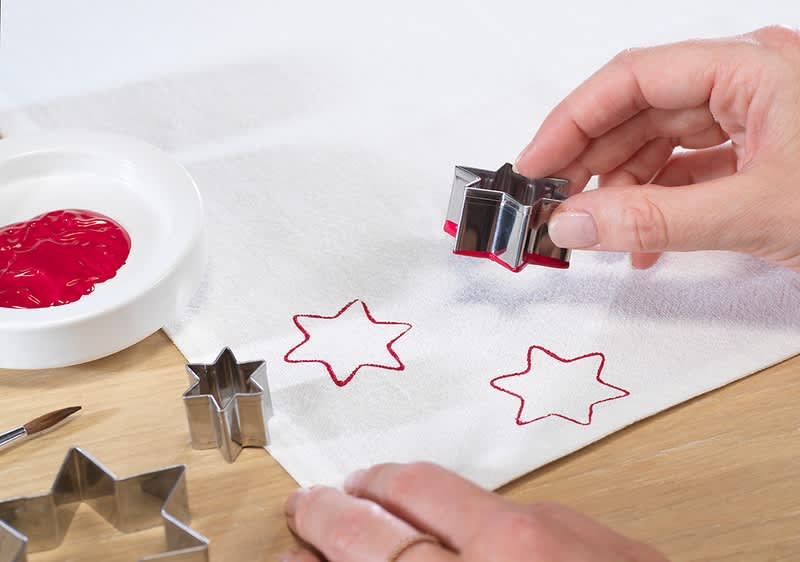 Eine Hand drückt einen sternförmigen Keksausstecher mit roter Farbe auf ein weißes Tuch, daneben ein kleiner Teller mit roter Farbe und ein Pinsel auf einem Holztisch