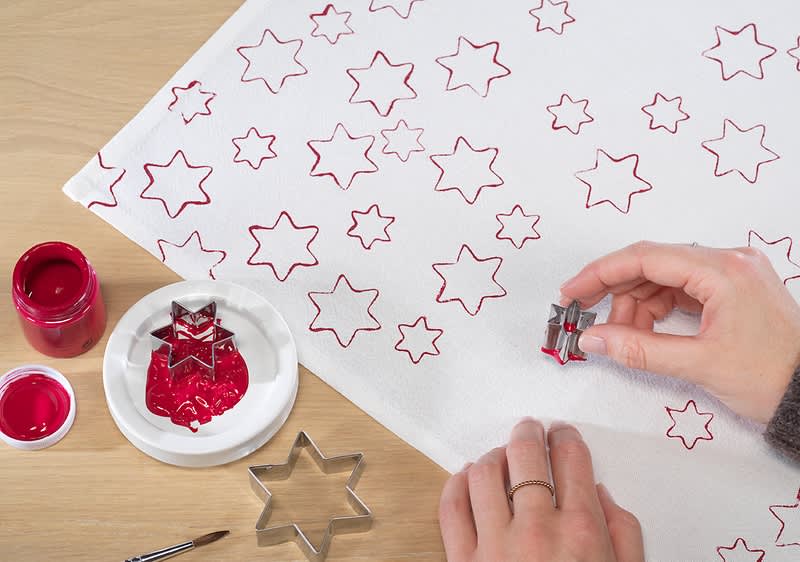 Eine Hand stempelt mit einem sternförmigen Keksausstecher rote Sterne auf ein weißes Tuch. Daneben stehen rote Farbe und weitere Ausstecher.