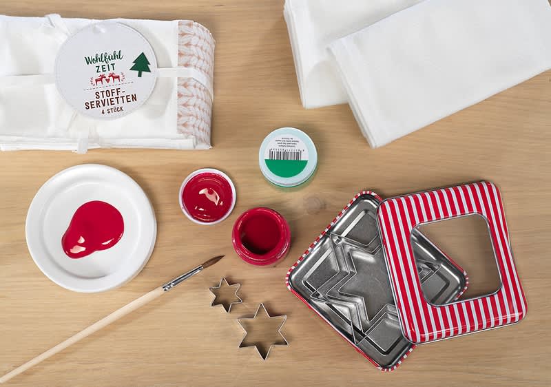 Stoff-Servietten-Set mit Textilfarbe, Pinsel und sternförmigen Keksausstechern auf einem Holztisch, daneben eine geöffnete rot-weiß gestreifte Dose