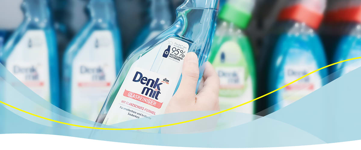 Eine Hand hält eine Flasche Denkmit Glasreiniger mit der Aufschrift Denk mit Glasreiniger mit Klarsicht-Formel und 95% weniger Plastik, im Hintergrund sind unscharf weitere Flaschen von Denkmit Reinigungsprodukten in verschiedenen Farben zu sehen