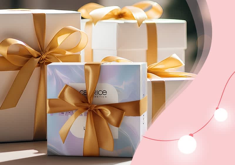 Drei weiße Geschenkboxen mit goldenen Schleifen, eine Box mit Catrice-Aufschrift und goldener Schleife, daneben rosa Hintergrund mit Lichterkette