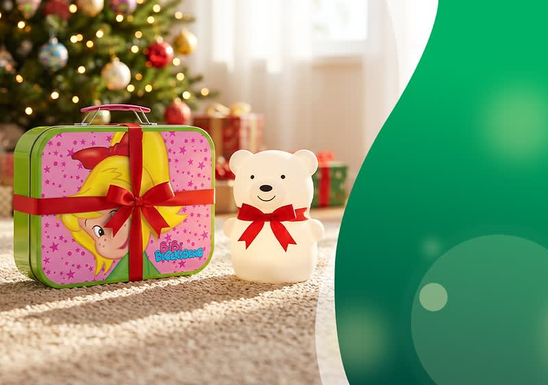 Rechteckige Geschenkbox mit Comic-Mädchen-Motiv, rot gebundenes Band daneben eine weiße Bärenfigur mit roter Schleife, im Hintergrund unscharfer Weihnachtsbaum und Geschenke. Bild mithilfe von KI erstellt